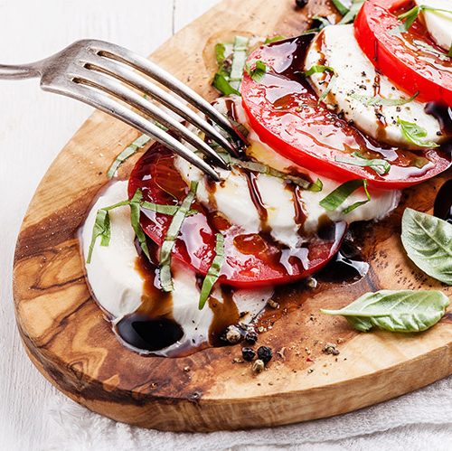 A caprese salad of sliced mozzarella, sliced tomatoes, basil, and balsamic, is plated on a wooden cutting board.